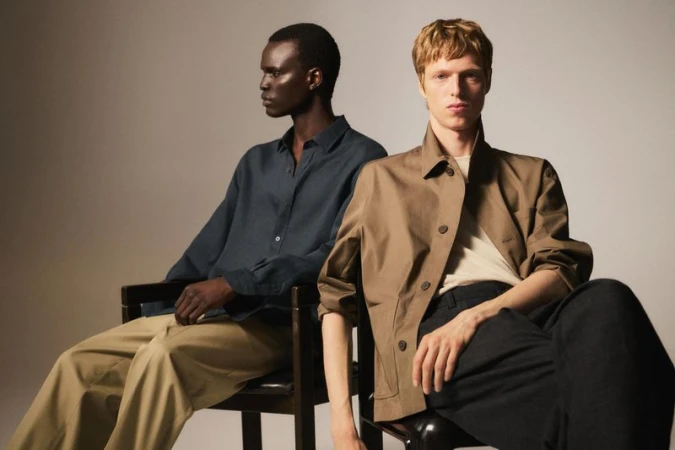 Two adult men sit indoors under studio lighting wearing casual fashion clothing in a portrait shot with a neutral background.