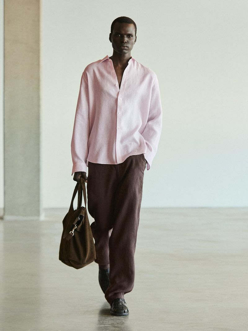 A dark-skinned man walks toward the viewer wearing a pink button-down shirt and dark pants while carrying a dark brown tote bag indoors.