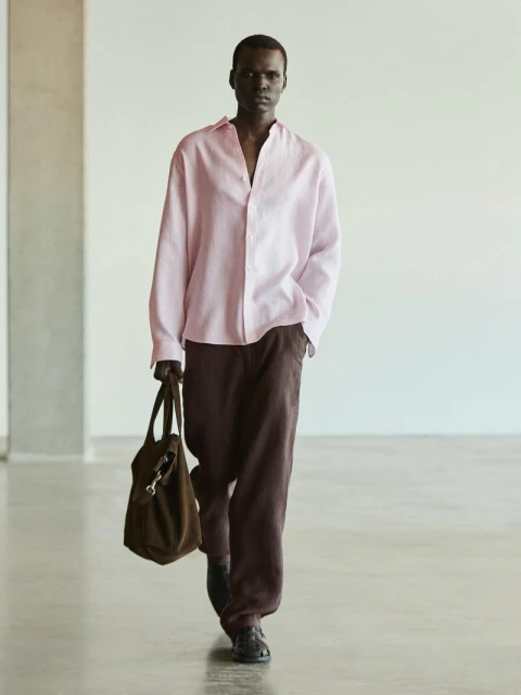 A dark-skinned man walks toward the viewer wearing a pink button-down shirt and dark pants while carrying a dark brown tote bag indoors.