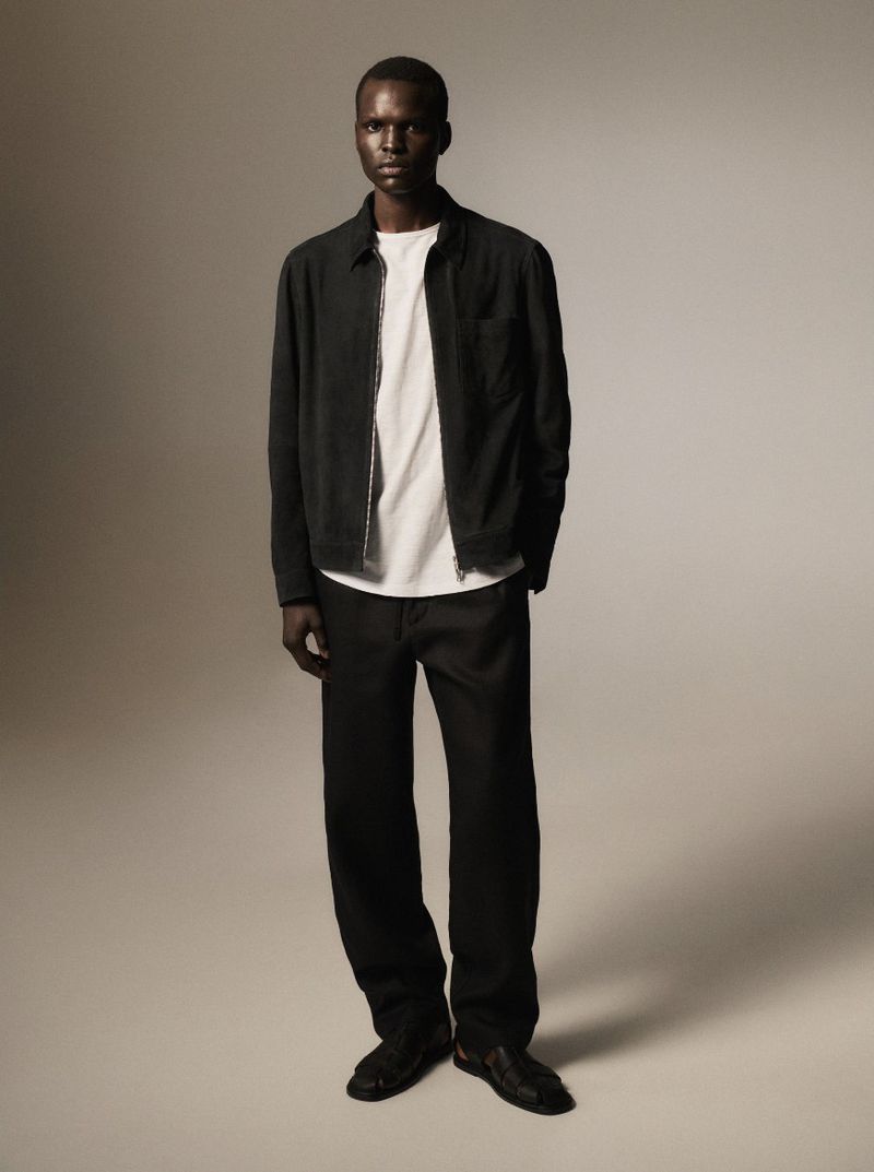 Full-length studio shot of an adult male fashion model wearing a black jacket, white shirt, and black pants against a grey background.