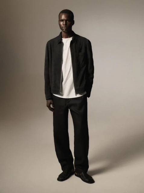Full-length studio shot of an adult male fashion model wearing a black jacket, white shirt, and black pants against a grey background.