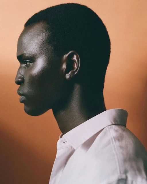 Portrait of a dark-skinned man in profile, wearing a white collared shirt against a soft orange background with shadow play.
