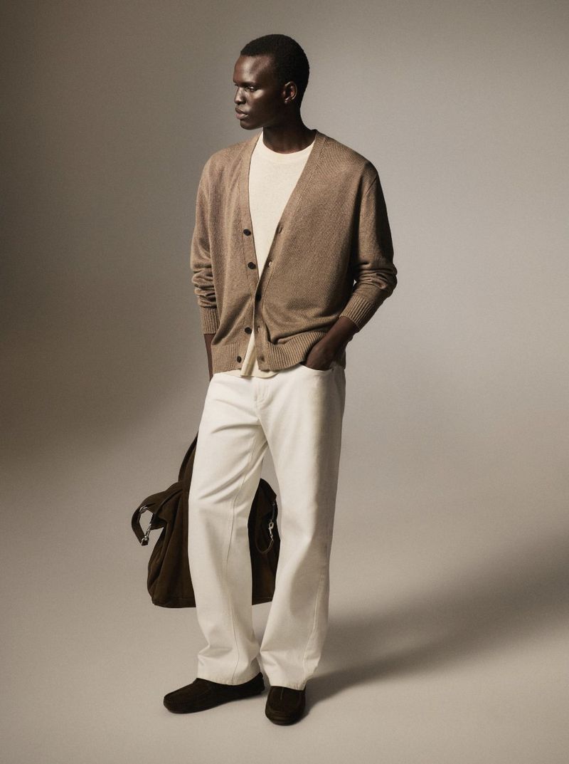 A stylish adult male model in an open brown cardigan, white shirt, and white denim trousers, standing in a studio with a shoulder bag.