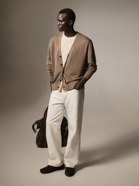 A stylish adult male model in an open brown cardigan, white shirt, and white denim trousers, standing in a studio with a shoulder bag.