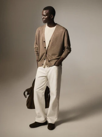 A stylish adult male model in an open brown cardigan, white shirt, and white denim trousers, standing in a studio with a shoulder bag.