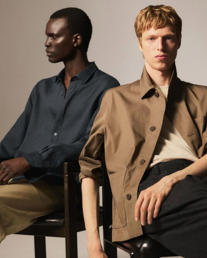 Two men in casual fashion, one African-American in a dark shirt, one fair-skinned in a tan jacket, sitting in a studio, neutral background.