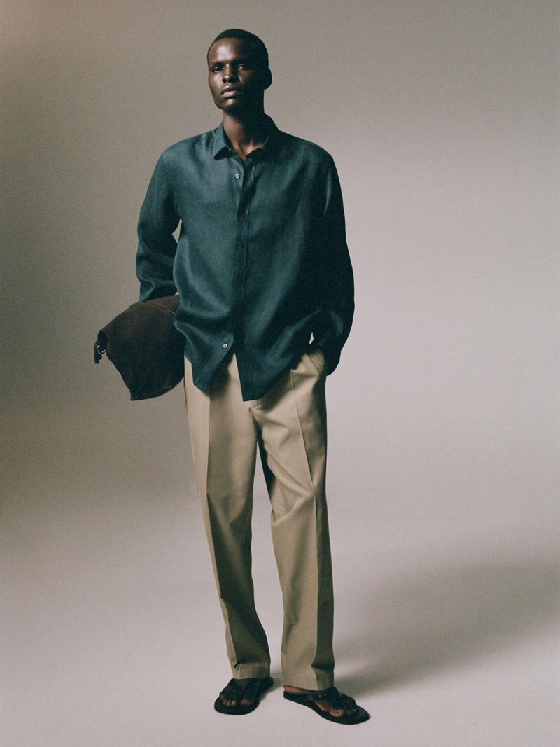 An adult male model poses indoors wearing a blue shirt, tan pants, and sandals. He is holding a bag over his shoulder.