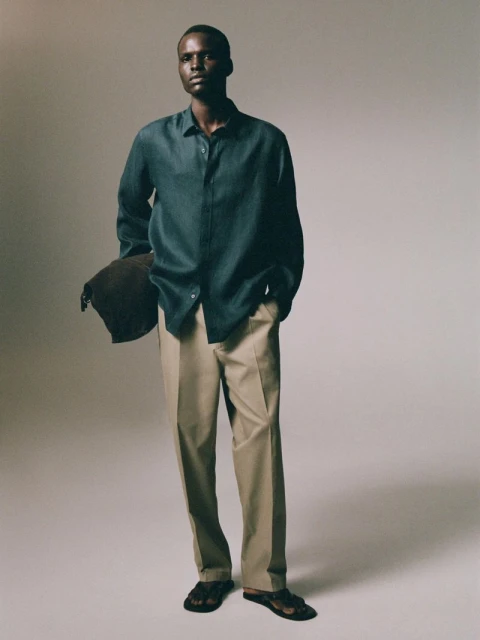 An adult male model poses indoors wearing a blue shirt, tan pants, and sandals. He is holding a bag over his shoulder.