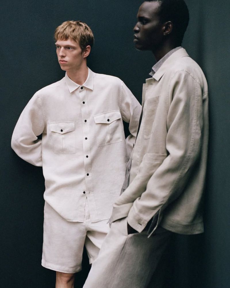 Two male models pose in matching beige linen button-down shirts and pants in a studio setting. The models are wearing casual attire.