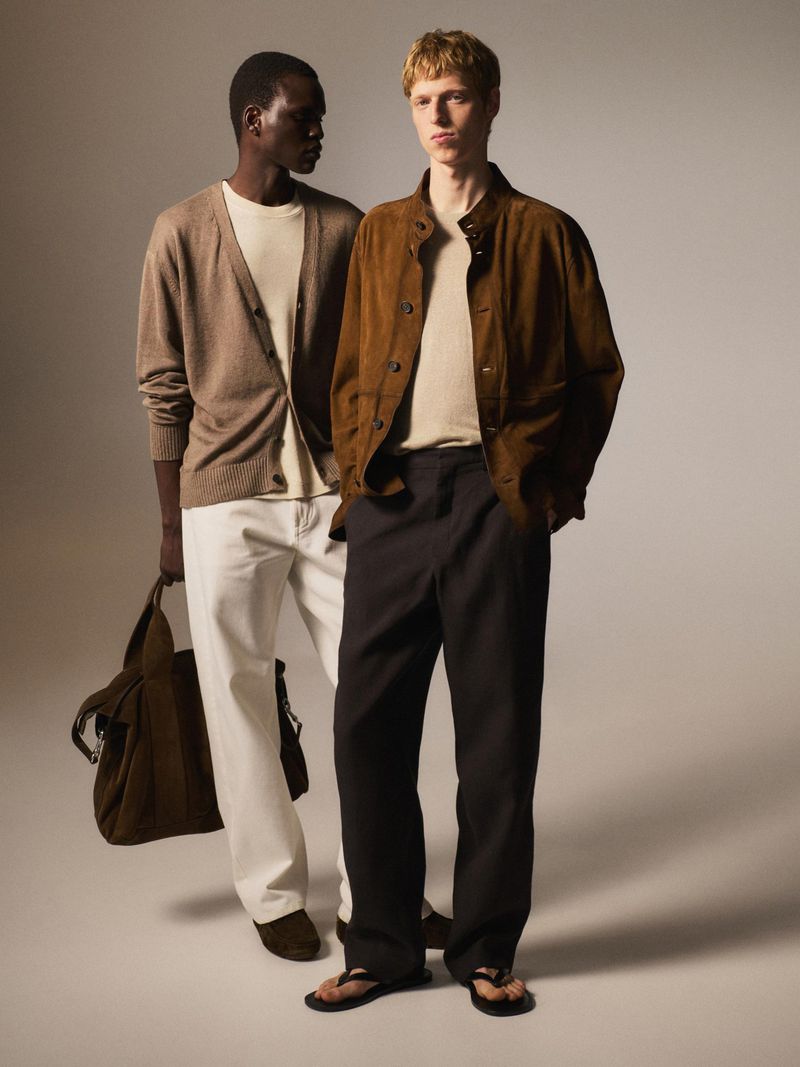 Two men in casual fashion pose in a studio. One carries a brown tote bag, wearing a cardigan and white pants. The other wears a suede jacket.