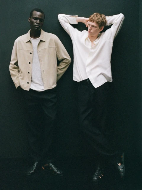 Two men pose in a studio wearing casual attire. One man wears a tan jacket with black pants and the other a white shirt with black pants.