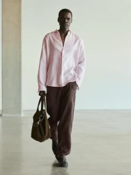 Black male model wearing a light pink long-sleeve shirt and dark trousers holding a brown suede tote bag in a minimalist interior