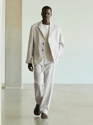 Black male model wearing an off-white blazer, light t-shirt, and trousers walking forward in a minimal studio setting.