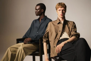 Two male models seated on chairs wearing relaxed summer shirts and trousers against a neutral studio backdrop.