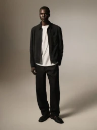 Model Badhiel Lony Nyang wearing a black zip jacket, white t-shirt, and black trousers against a warm neutral studio backdrop.
