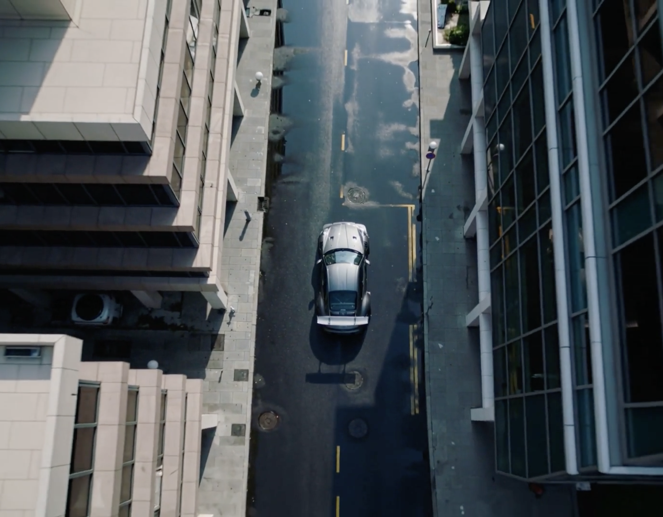 Aerial view of a silver sports car with a large rear wing driving down a wet city street between modern commercial buildings.