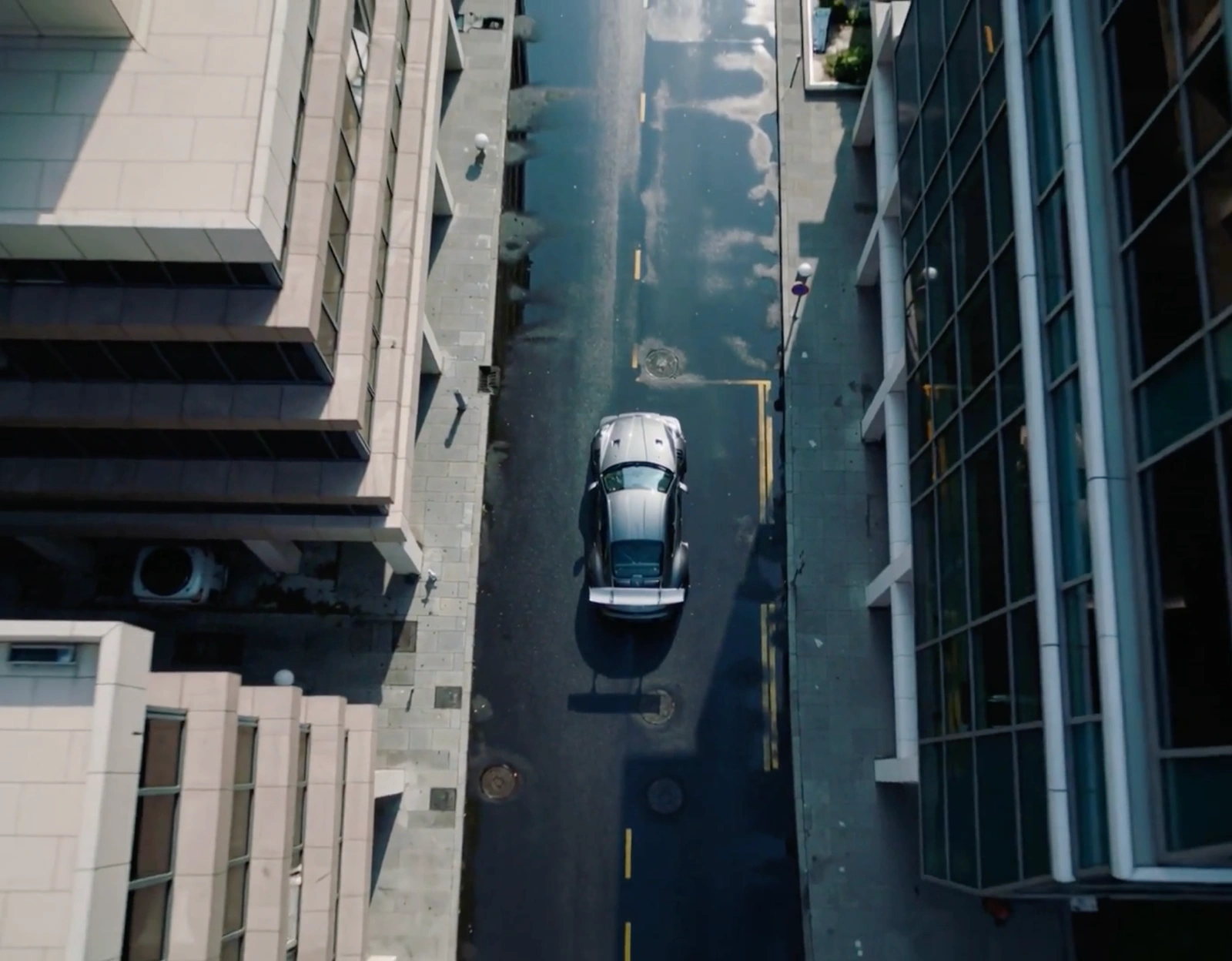 Aerial view of a silver sports car with a large rear wing driving down a wet city street between modern commercial buildings.