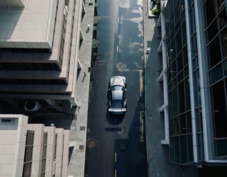 Aerial view of a silver sports car with a large rear wing driving down a wet city street between modern commercial buildings.