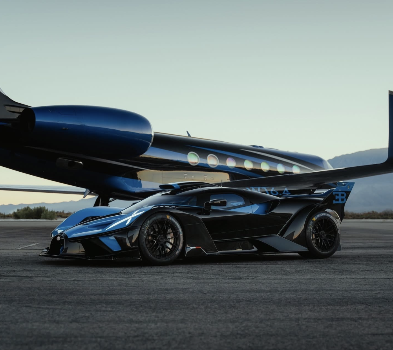 A blue and black Bugatti hypercar parked on an airport tarmac in front of a dark luxury private jet with mountains in the background.