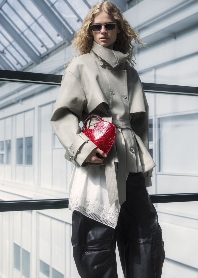 A stylish blonde woman wearing a trench coat and sunglasses holds a red heart-shaped purse against a bright indoor background.