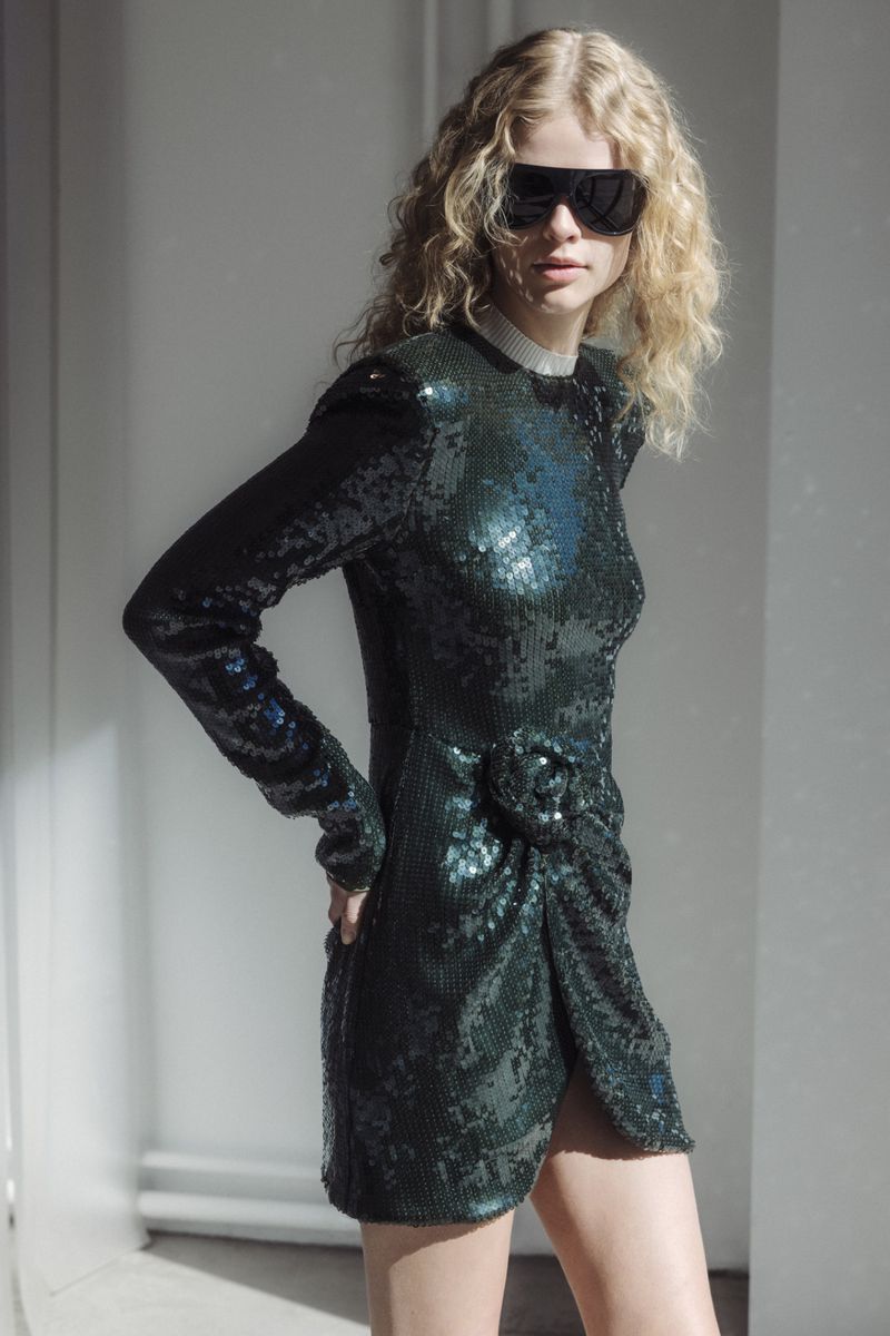 Model wearing a dark green sequin long-sleeve mini dress and black shield sunglasses against a bright wall.