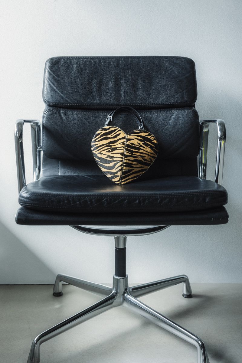 Heart-shaped animal print handbag placed on a black leather executive office chair against a plain light wall.