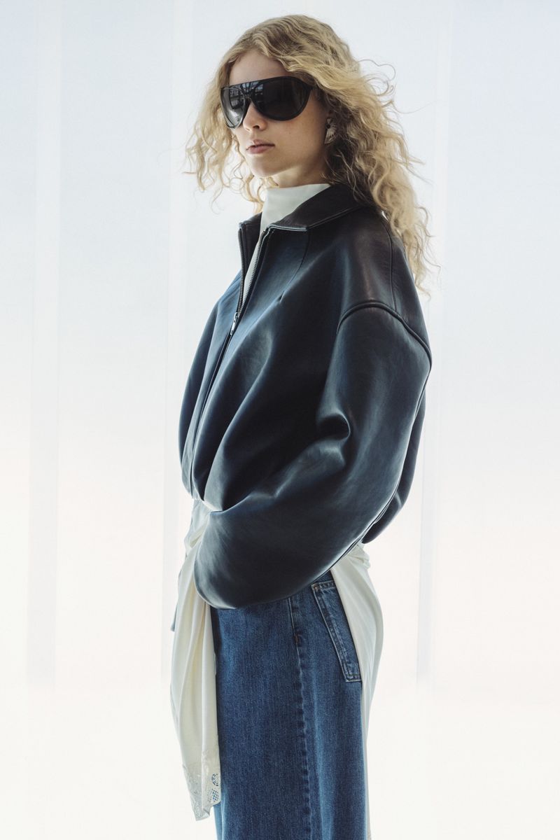 Model wearing an oversized black leather jacket, shield sunglasses, and blue denim jeans against a bright white background.