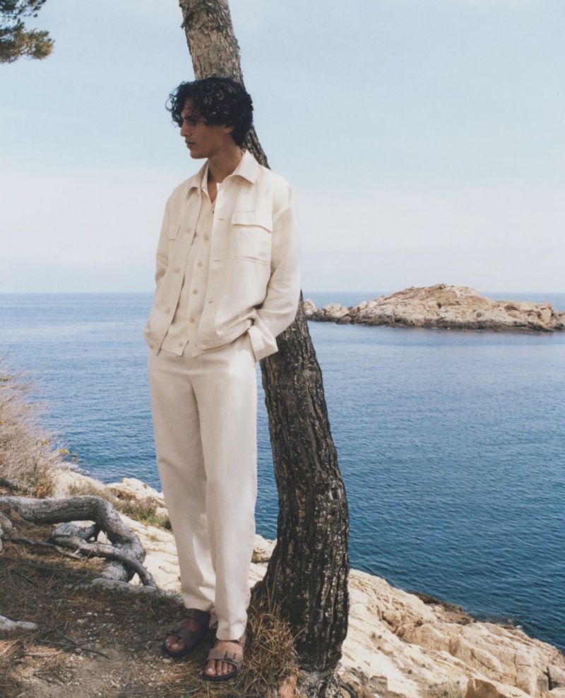 A relaxed young man leans against a tree in a light-colored linen suit with the ocean and a rocky island visible in the background.