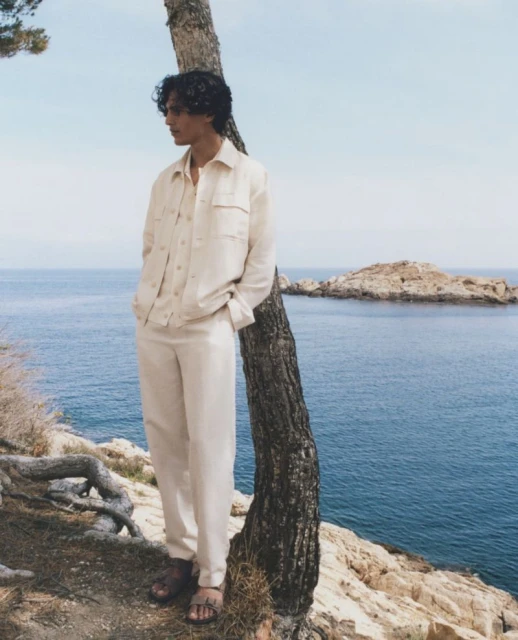 A relaxed young man leans against a tree in a light-colored linen suit with the ocean and a rocky island visible in the background.