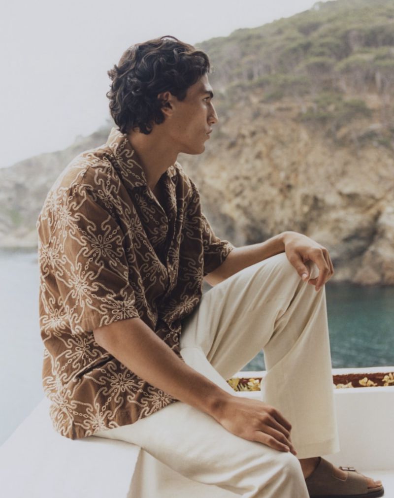 Profile shot of a young adult male model in a patterned brown shirt and cream-colored pants, sitting and looking out to a sea and mountain view.