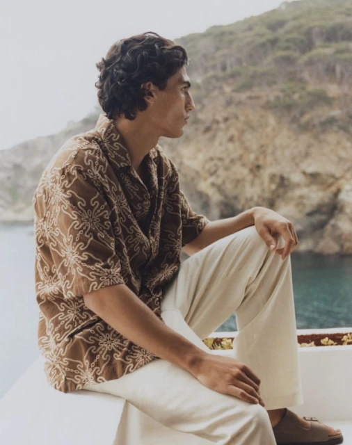 Profile shot of a young adult male model in a patterned brown shirt and cream-colored pants, sitting and looking out to a sea and mountain view.
