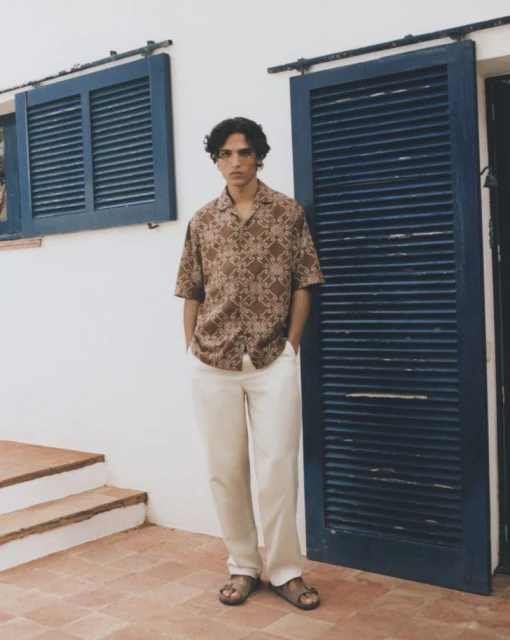 A young adult man poses outdoors in patterned shirt, light trousers, and sandals near a blue shuttered window and white wall with terracotta steps.