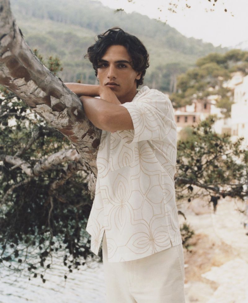 Man in patterned shirt leans against tree outdoors. Portrait showcases casual fashion in a natural setting with mountains in the background.