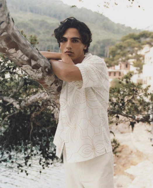 Man in patterned shirt leans against tree outdoors. Portrait showcases casual fashion in a natural setting with mountains in the background.