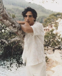 Model leaning against a tree branch wearing a patterned white short sleeve shirt in an outdoor coastal setting