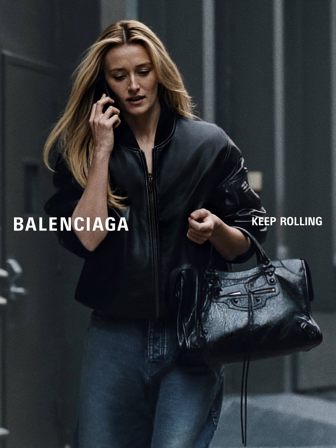 Sarah Pidgeon walking in a black leather bomber jacket and jeans while holding a black Balenciaga handbag and talking on a smartphone.