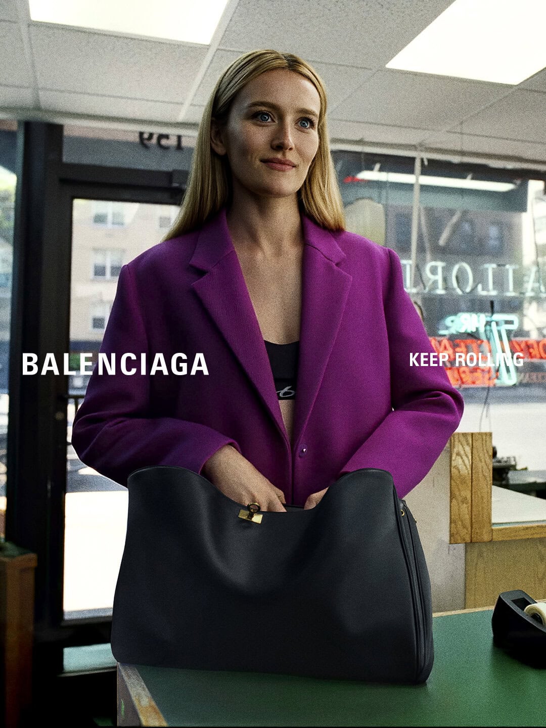 Sarah Pidgeon in a magenta wool blazer opening a large black leather tote bag at a tailor shop counter.