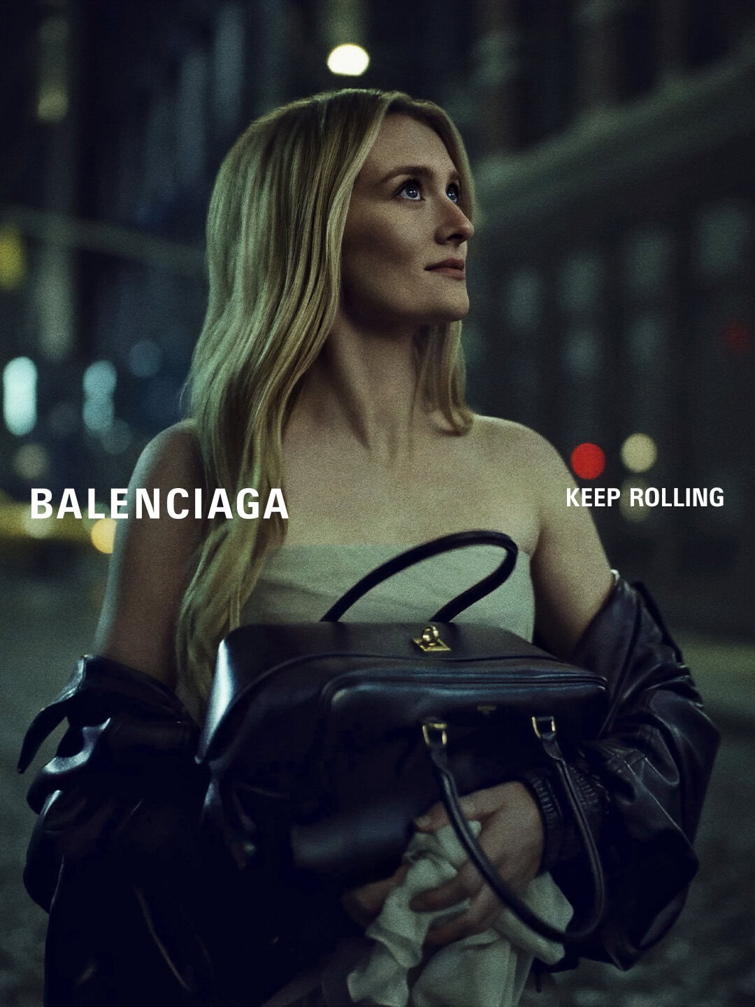 Sarah Pidgeon holding a black leather handbag and wearing a strapless top with a dark jacket at night on a city street.