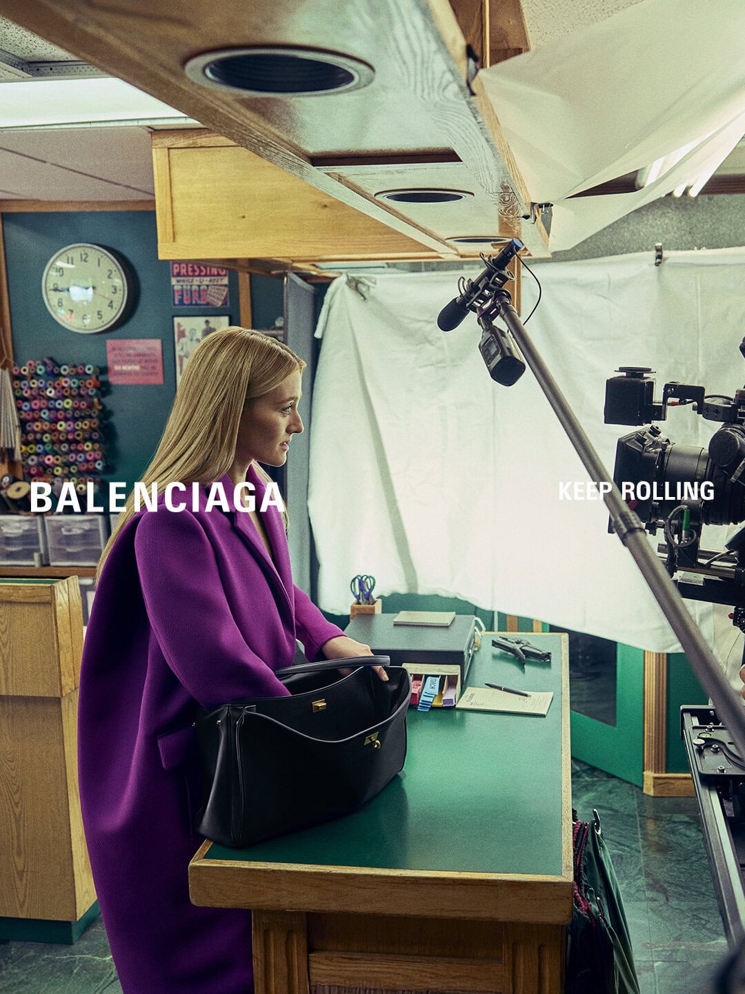 Sarah Pidgeon wearing a bold purple coat beside a large black leather handbag on a shop counter with filming equipment.