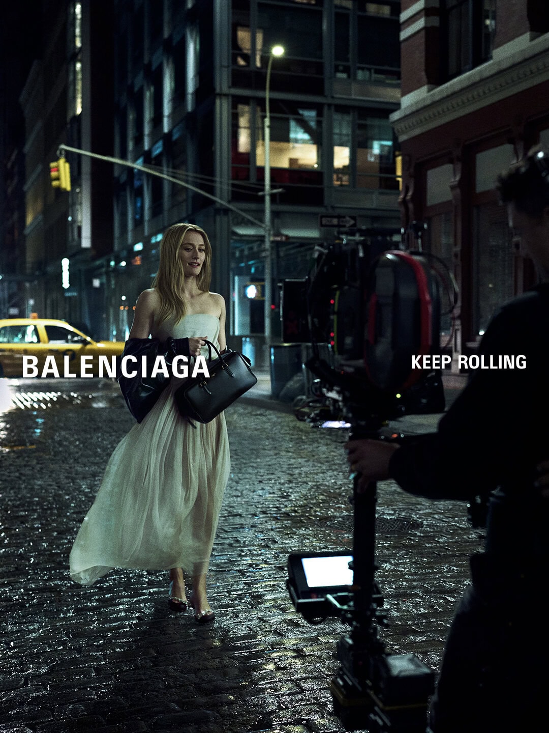 Fashion photoshoot on a city street at night featuring a woman in a dress with handbag, Balenciaga logo, and film crew.