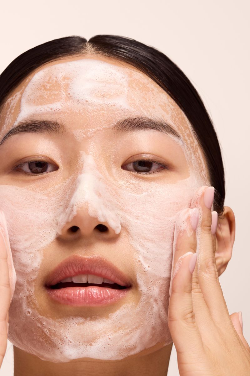 Close-up portrait of an East Asian woman applying a white foaming face cleanser across her cheeks and forehead with both hands.