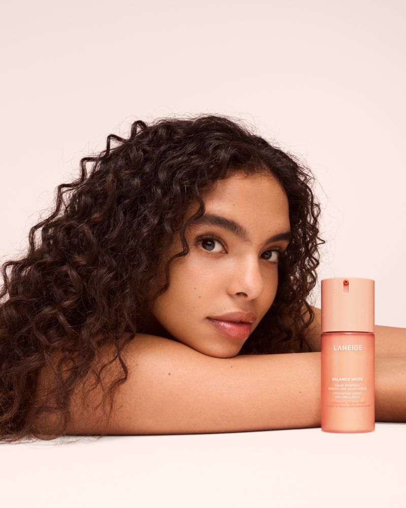 Woman with dark curly hair resting her chin on her arm next to a Laneige skincare bottle against a neutral studio backdrop.