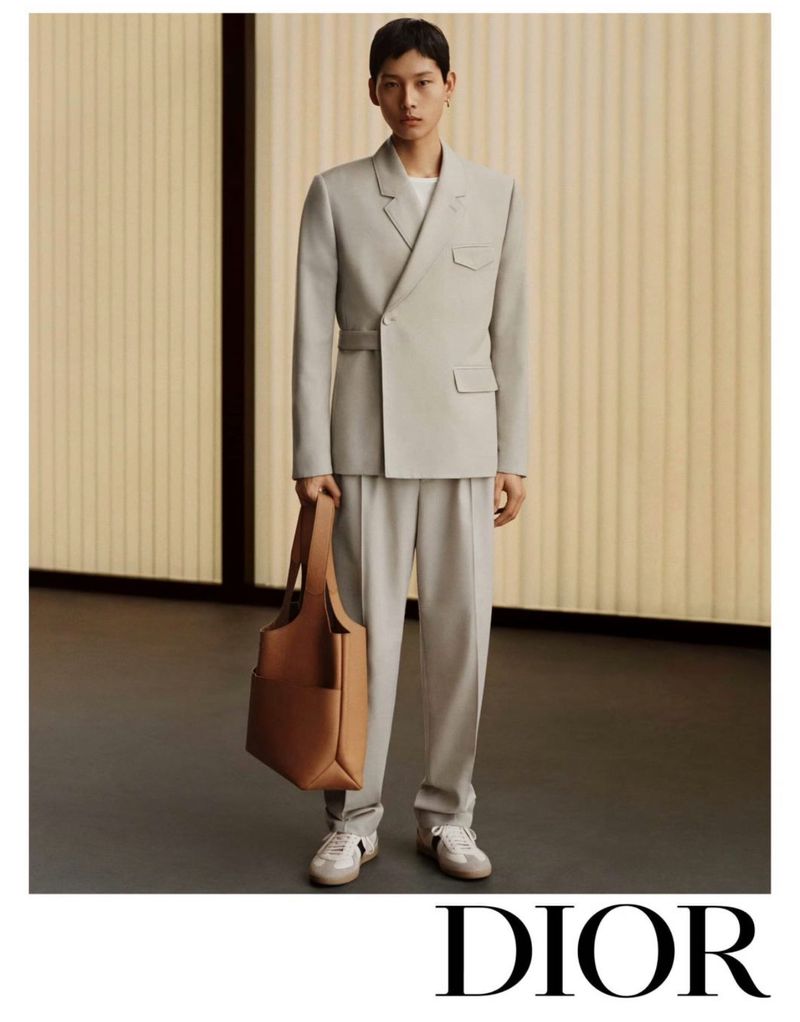 A young Asian man models a pale grey suit with a tote bag in a fashion-forward studio portrait for a Dior advertisement.