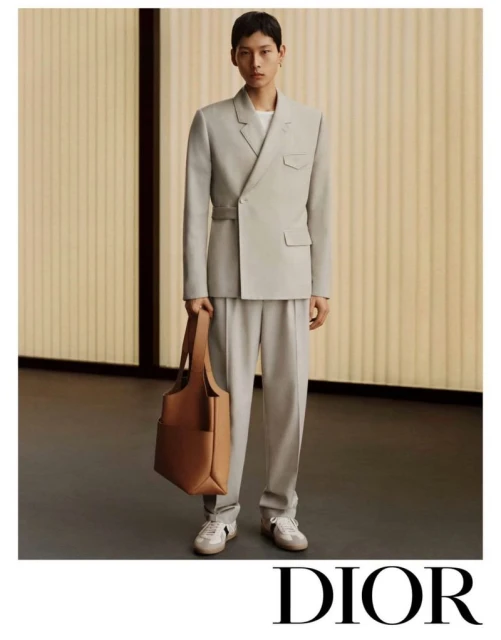 A young Asian man models a pale grey suit with a tote bag in a fashion-forward studio portrait for a Dior advertisement.