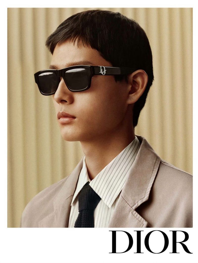 A male model in a light tan suit and dark sunglasses poses against a neutral background in a fashion editorial shot by DIOR.