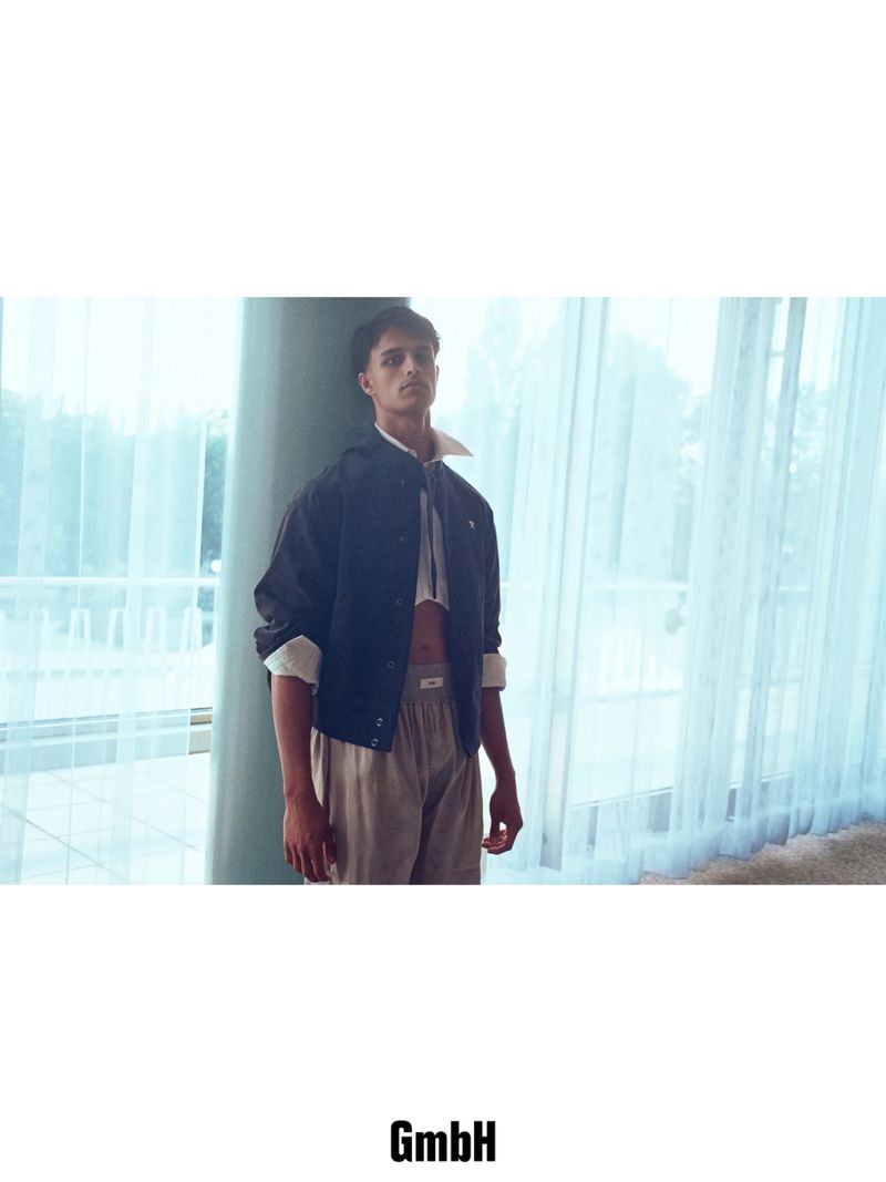 Model in a dark jacket, cropped white shirt, and beige trousers standing by sheer curtains for GmbH Spring 2026.