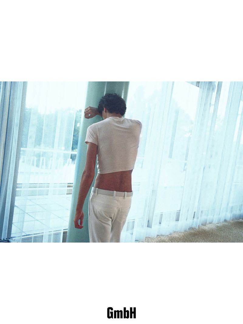 Model with back turned in a cropped t-shirt and light trousers leaning against a column before sheer window curtains.