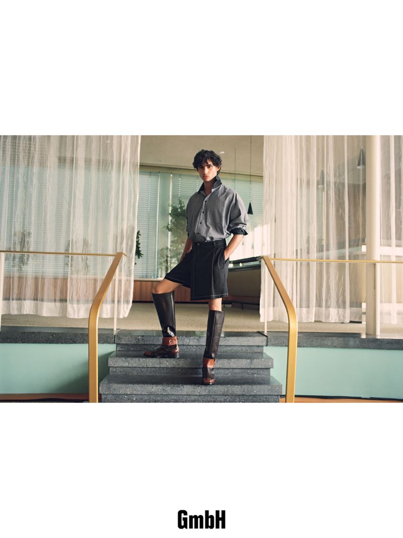 Model wearing a grey collared shirt, dark shorts, and two-tone knee-high riding boots standing on interior stone steps.