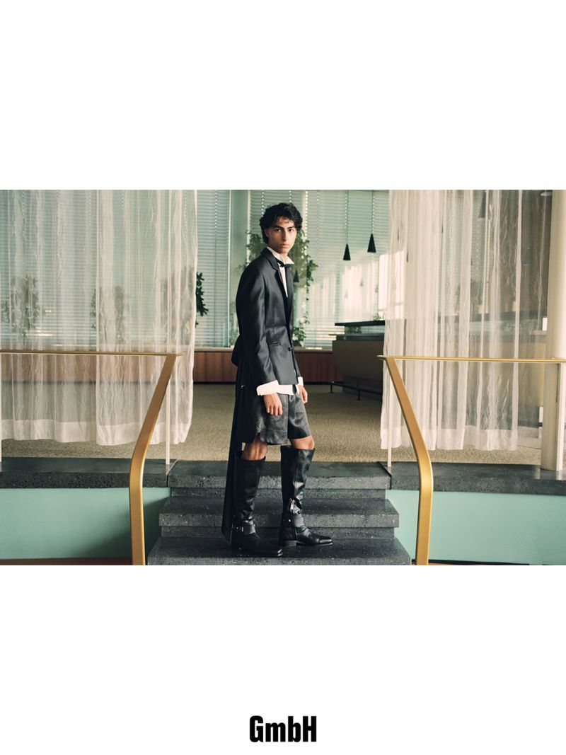 A person in a suit, shorts, and boots stands on a set of stairs inside a building with floor-to-ceiling window coverings and sparse decor.