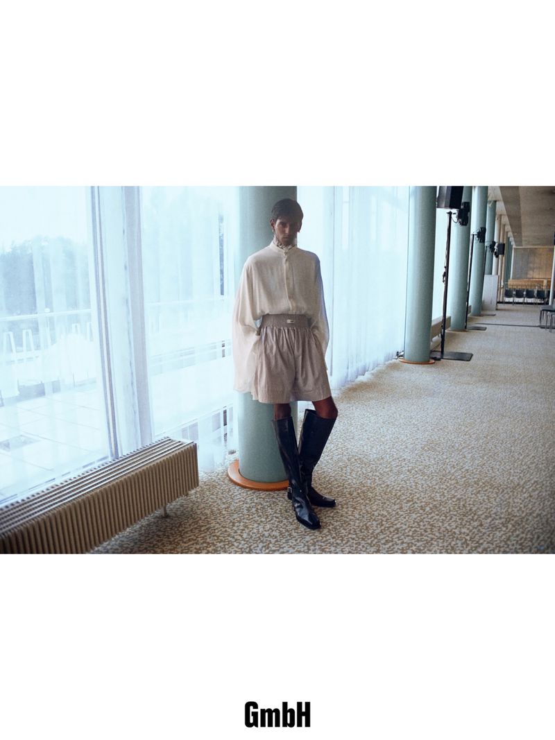 Model wearing a draped cream blouse, belted shorts, and tall black leather boots standing by a pillar near large windows.